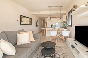 Ένα μοντέρνο σαλόνι με έναν γκρι καναπέ, ένα τραπεζάκι σαλονιού, μια τηλεόραση επίπεδης οθόνης και μια τραπεζαρία με μια κουζίνα στο βάθος.