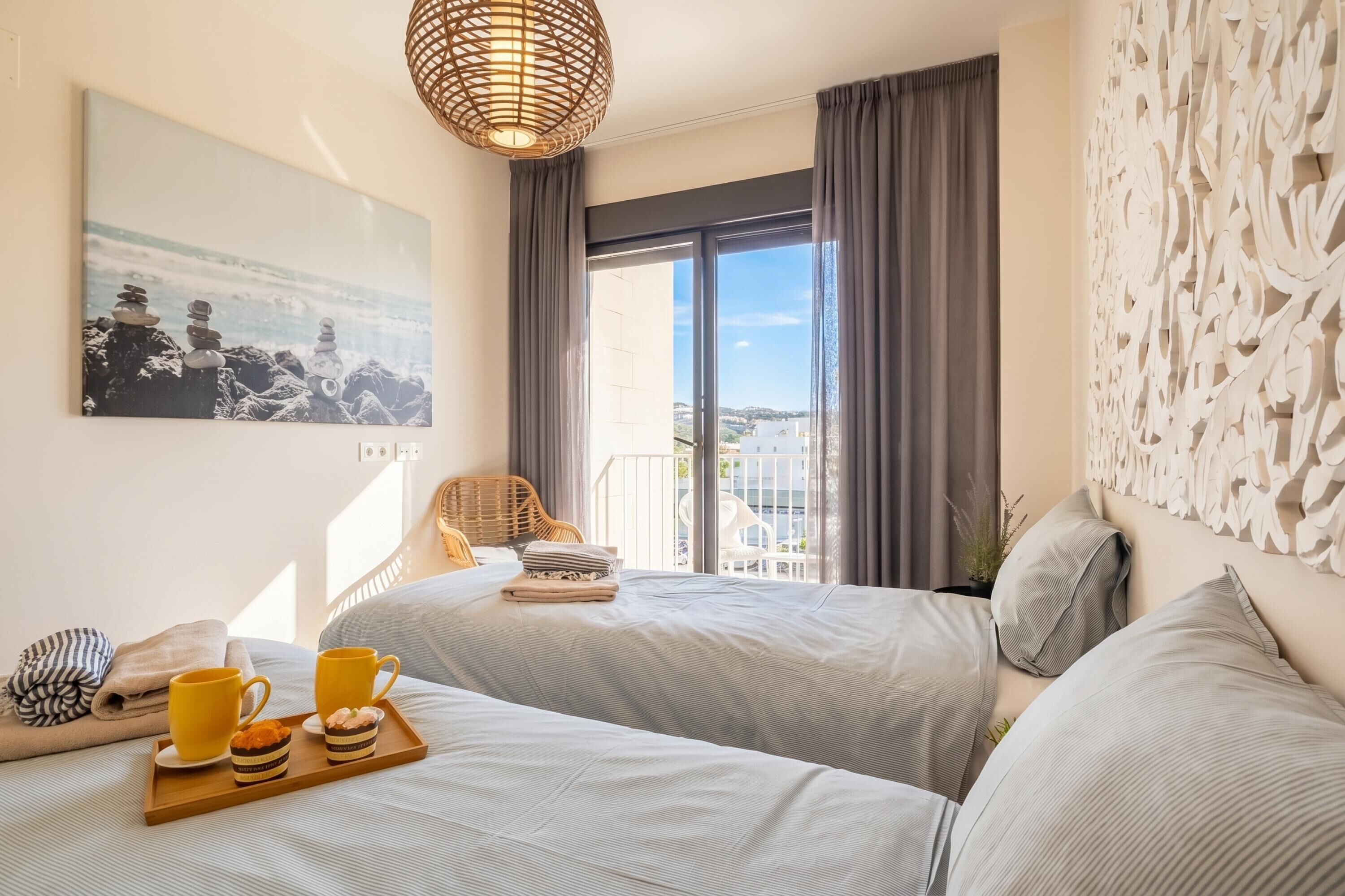 Hotelkamer met twee bedden, een eettafel met gele mokken, een rieten stoel, een balkon met uitzicht en een decoratief paneel aan de muur.