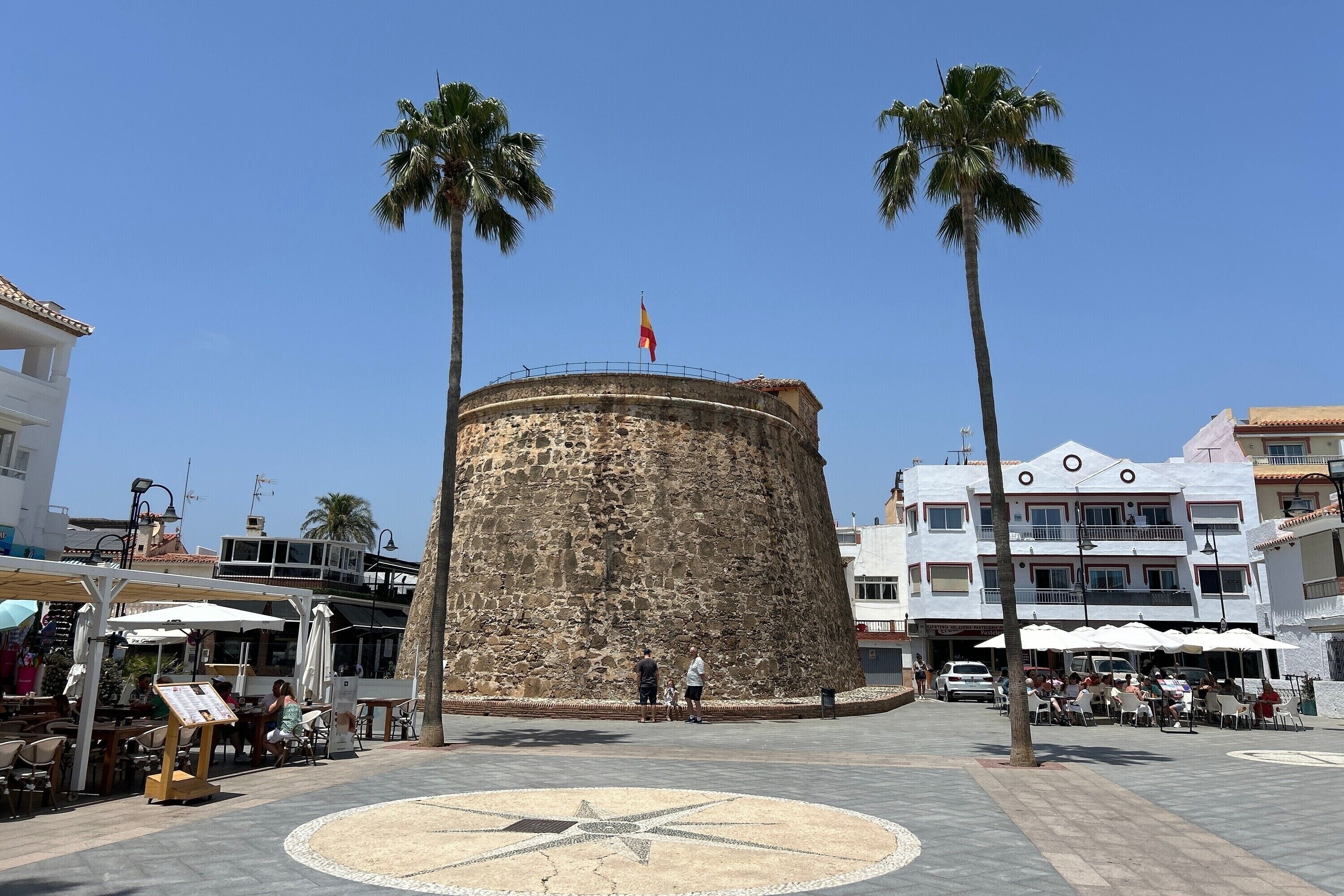 Una plaza con una torre de piedra, palmeras y zonas de estar al aire libre.