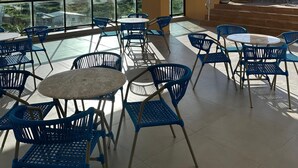 A covered outdoor seating area with blue chairs and tables, a glass balustrade, and a view of palm trees.