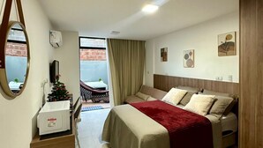 A hotel room with a bed, a small desk with a microwave, a chair, a window with curtains, and a view of a brick building outside.