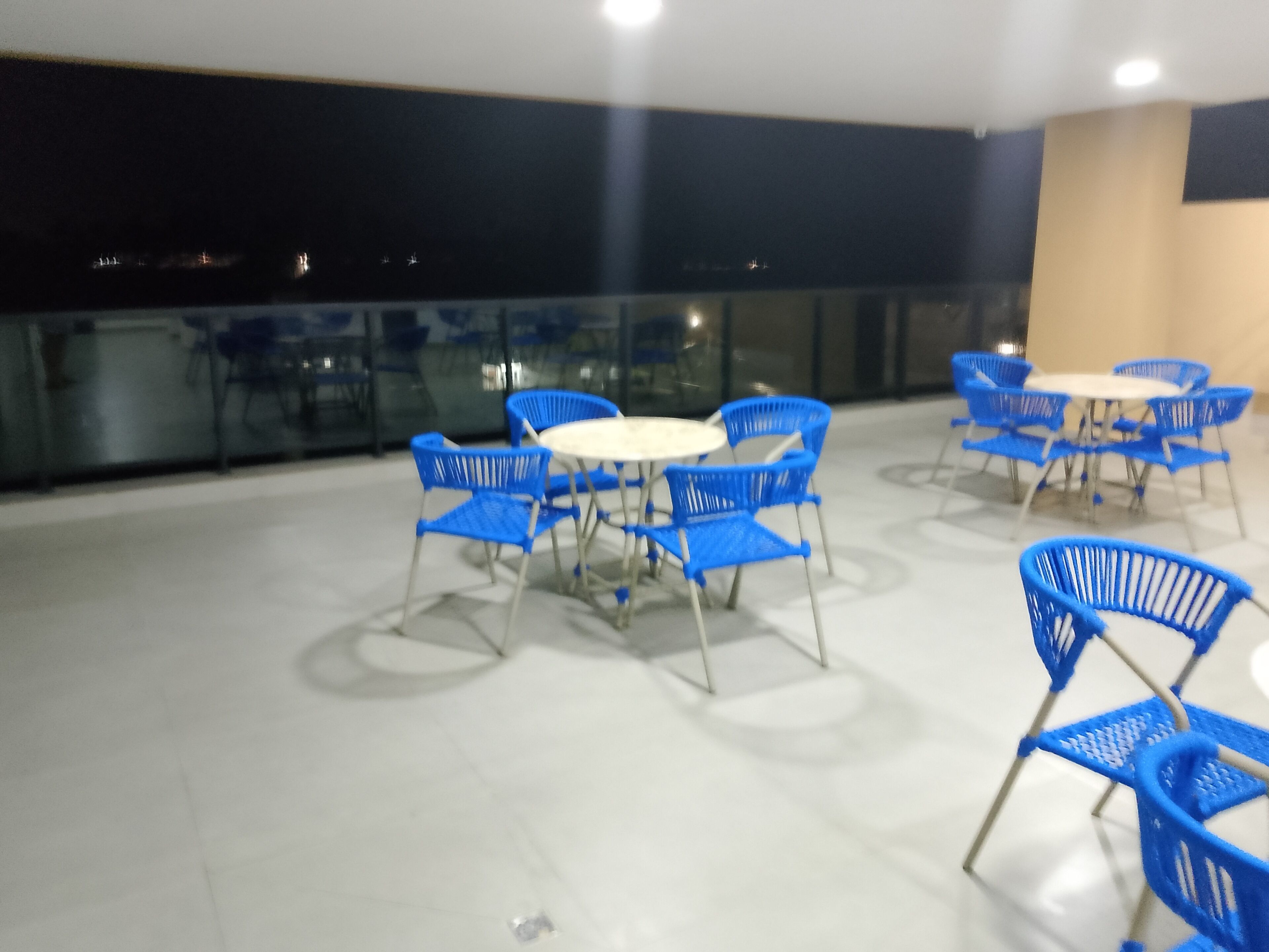 Une terrasse sur le toit, avec des chaises et des tables en plastique bleu, des rampes en verre et une vue sur la ville la nuit.