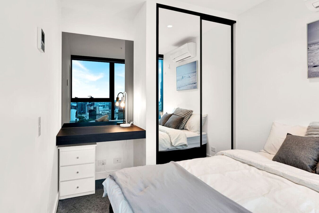 A modern bedroom with a city view, a desk with a lamp, a bed with pillows, and a large mirror.