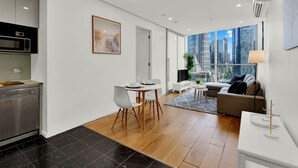 A modern apartment with a kitchen, dining area, and living room. It features a city view through large windows, a flat-screen TV, and a dining table with chairs.