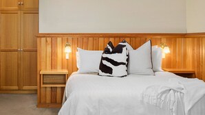 Una cama bien tendida con ropa blanca y dos almohadones decorativos, con mesitas de noche de madera y lámparas de pared.