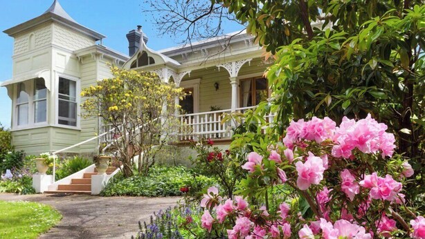 Una casa histórica con una torre destacada, un porche delantero y un jardín bien cuidado con flores rosadas.