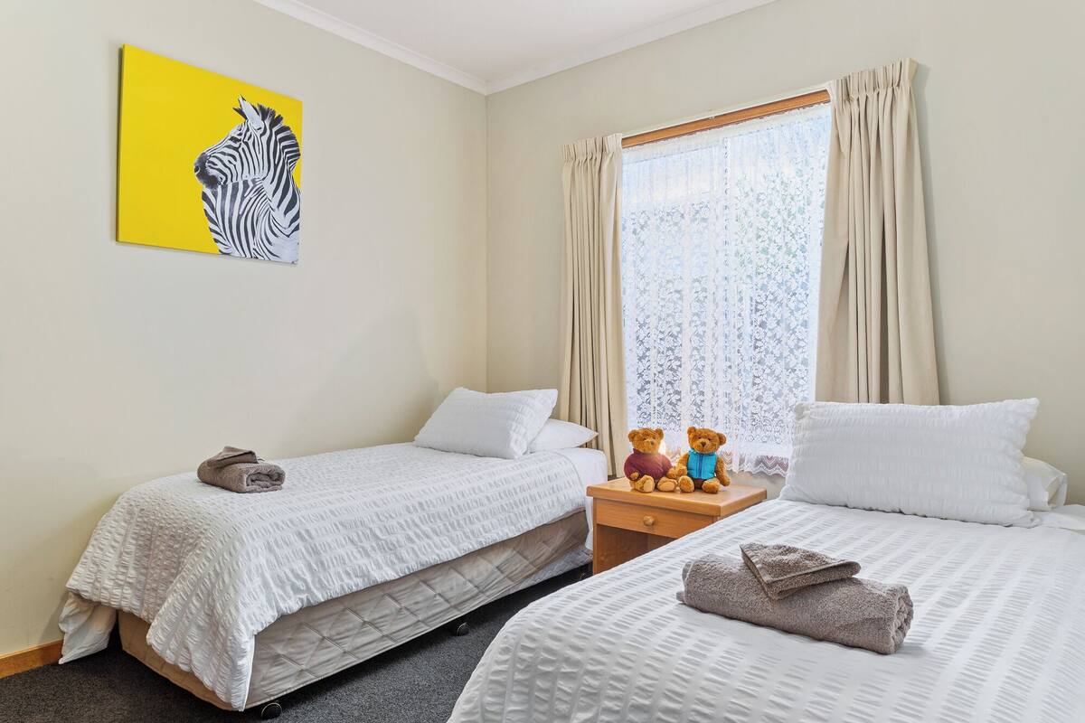 Ein Hotelzimmer mit zwei Betten, einem Fenster mit Vorhängen, einem Teddy auf dem Nachttisch und einem Zebra-Gemälde an der Wand.