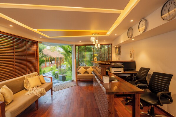 A modern living room with a wooden coffee table, sofa, and chairs. There is a television and a desk with a computer. The room has large windows and a view of greenery outside. - Alaya Stays Gracias Seaora Retreat (Morjim)