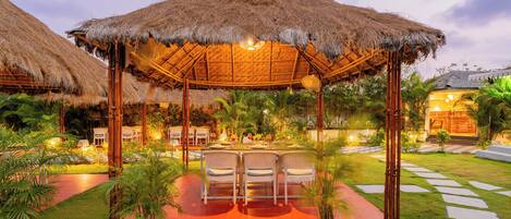 A thatched-roof outdoor dining area with chairs and tables, surrounded by potted plants and a well-maintained lawn.