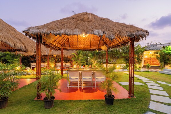 A thatched-roof outdoor dining area with chairs and tables, surrounded by potted plants and a well-maintained lawn. - Alaya Stays Gracias Seaora Retreat (Morjim)