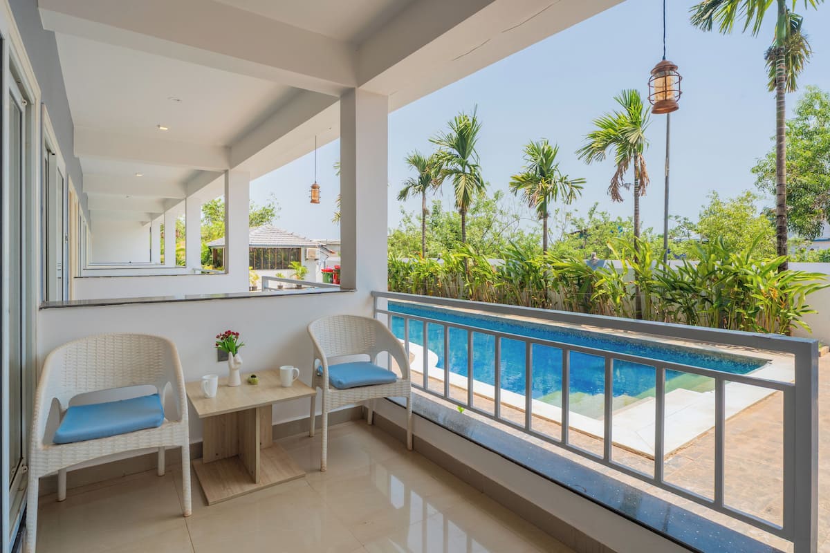 A balcony with a pool, wicker chairs, a table, and a view of palm trees and greenery.