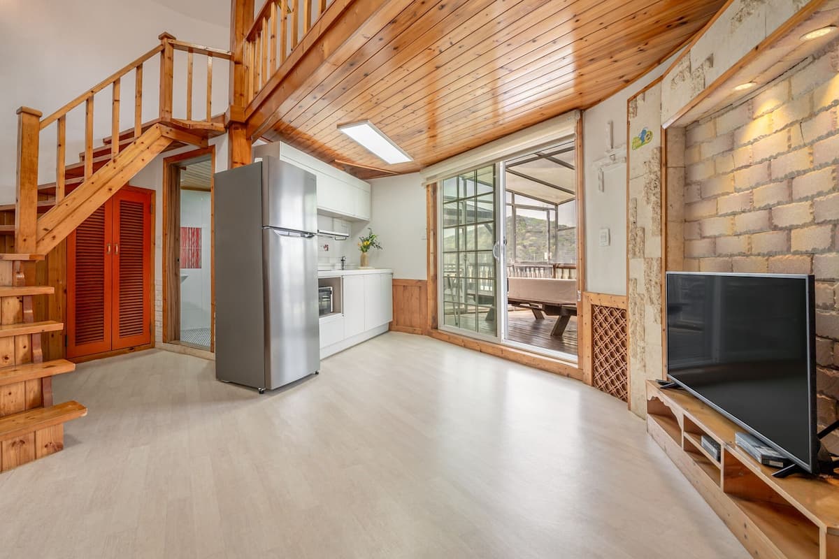 A spacious living area with a wooden staircase, a kitchen with a refrigerator, and a large TV on a wooden stand.