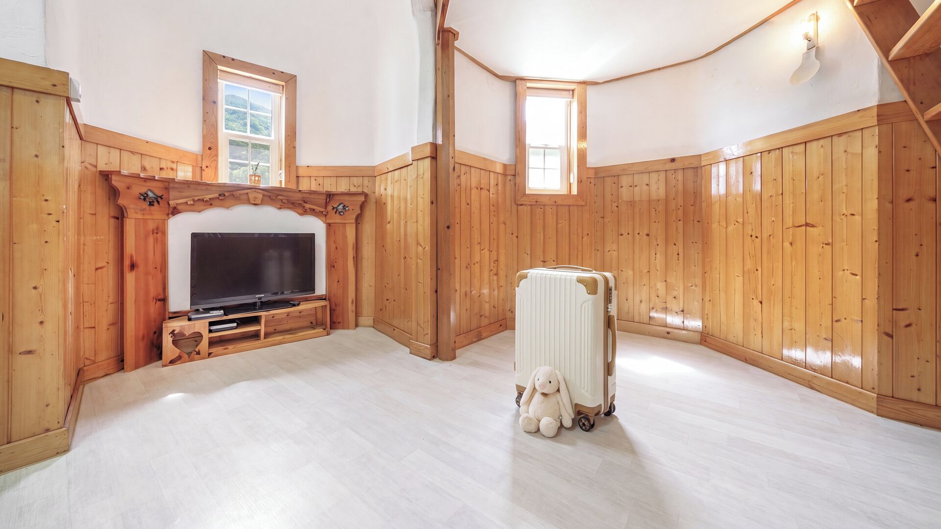 Une pièce aux murs en bois, un téléviseur posé sur un meuble en bois, une valise et un petit animal en peluche.