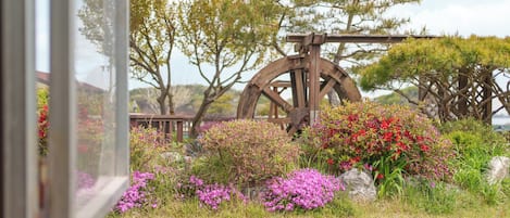 Un entorno rústico al aire libre con una gran noria de madera, rodeado de abundante vegetación y plantas en flor.