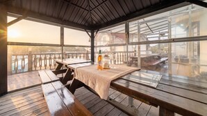 Een terras buiten met houten banken en picknicktafels, een fles drank en een glas op een geweven mat.