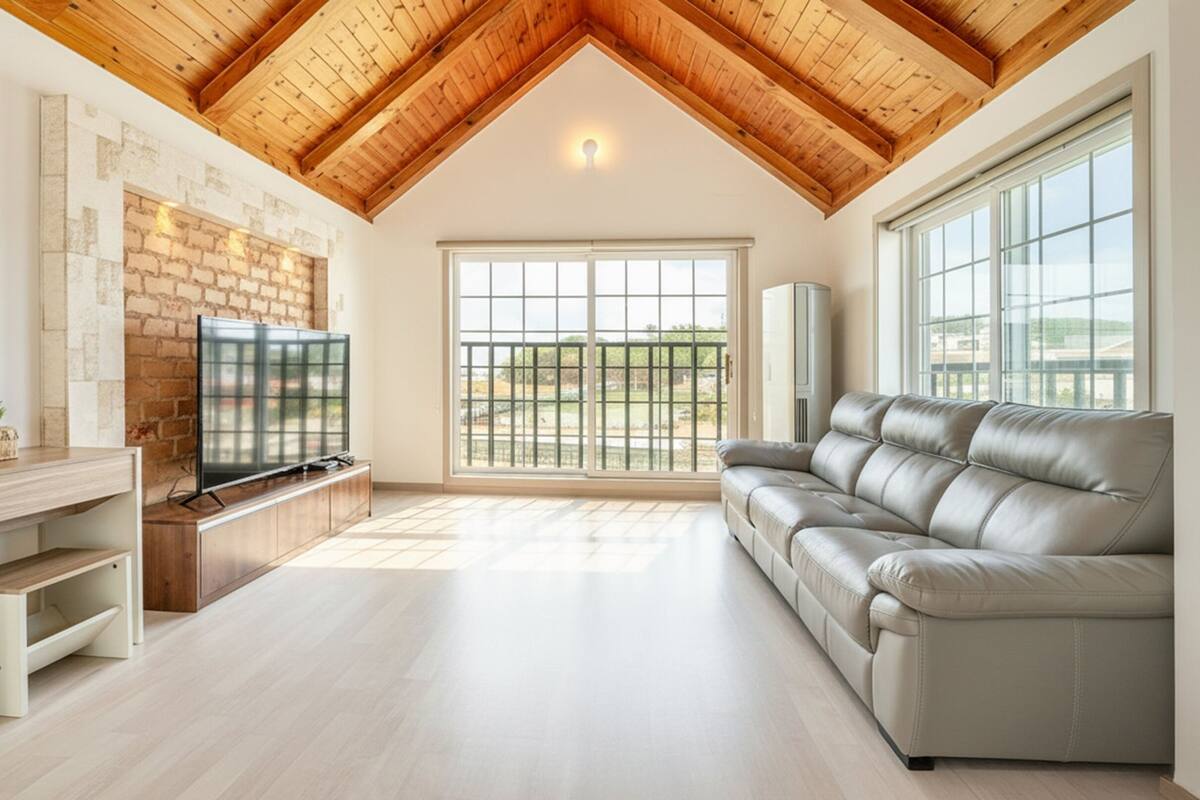 A spacious living room with a wooden ceiling, a large window, a leather sofa, and a flat-screen TV on a wooden stand.