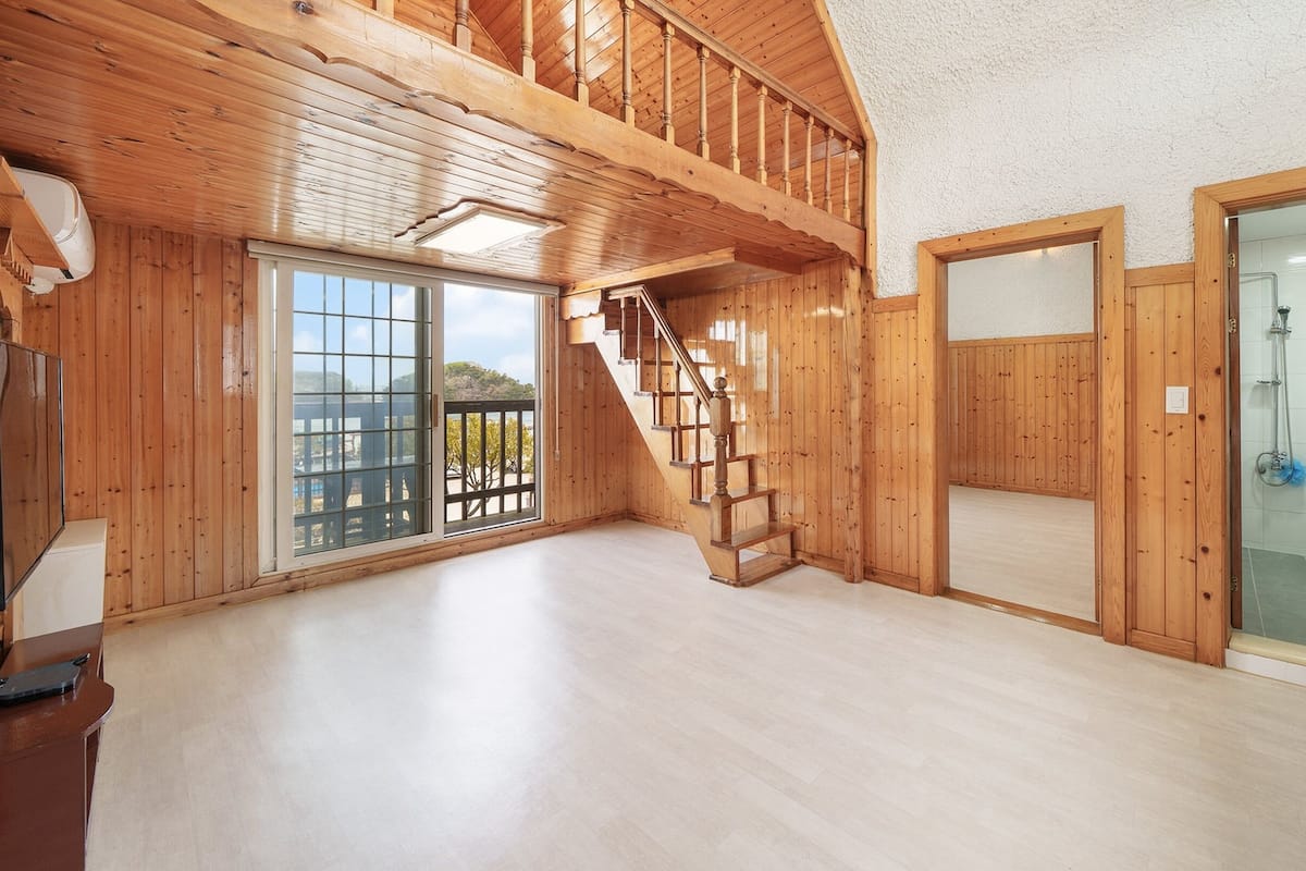 A spacious room with wooden walls, a wooden staircase, a glass shower, and a large window offering a view of the outdoors.