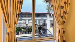 A window with curtains, a view of a traditional building with a tiled roof, and potted plants outside. - Hotel Thamla Haveli (Udaipur)
