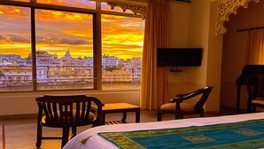 A hotel room with a large window offering a city view, a bed with a green and blue bedspread, two wooden chairs, and a flat-screen TV mounted on the wall. - Hotel Thamla Haveli (Udaipur)