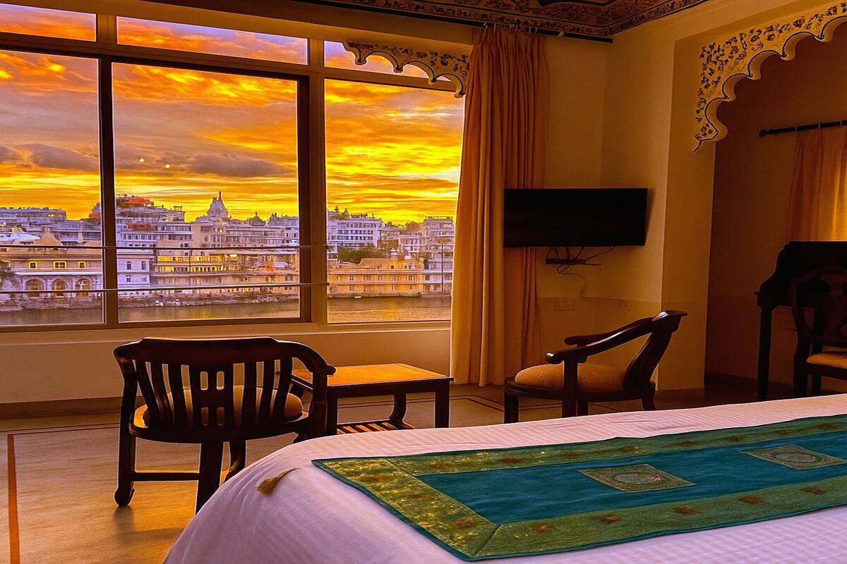 A hotel room with a large window offering a city view, a bed with a green and blue bedspread, two wooden chairs, and a flat-screen TV mounted on the wall.
