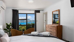 Een hotelkamer met een bed, twee stoelen, een houten dressoir, een spiegel en een raam met uitzicht op zee en bergen.