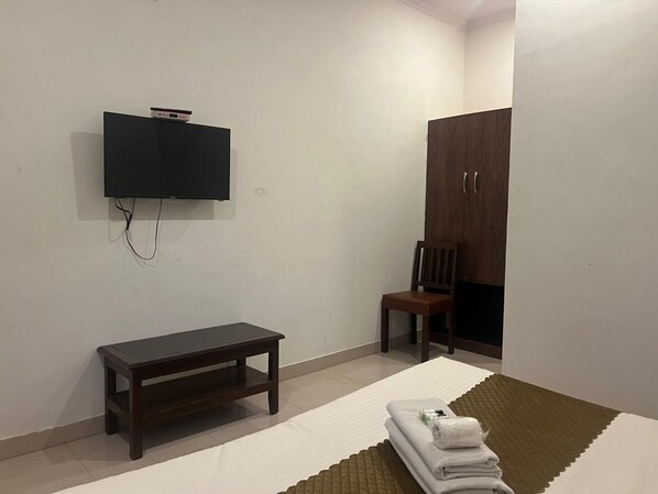 A room with a flat-screen TV mounted on the wall, a wooden bench, a wooden wardrobe, and a folded towel on the floor. - Akhey Vilas (Mount Abu)