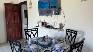 A dining area with a round table set for four, surrounded by chairs. A flat-screen TV is mounted on the wall above the table. - Glen Sea Inn of St. Mary - Charming 2 Bedroom Beachside Home (Port Maria)