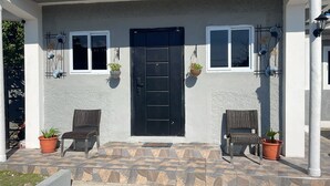 Une maison avec une porte noire, deux chaises en osier et des plantes en pot sur le porche.