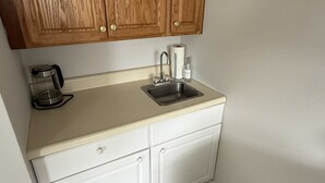 A small kitchen with wooden cabinets, a sink, a coffee maker, and a roll of paper towels. - Keel at Soundview · Beach+OceanView+Sunrise (Old Lyme)