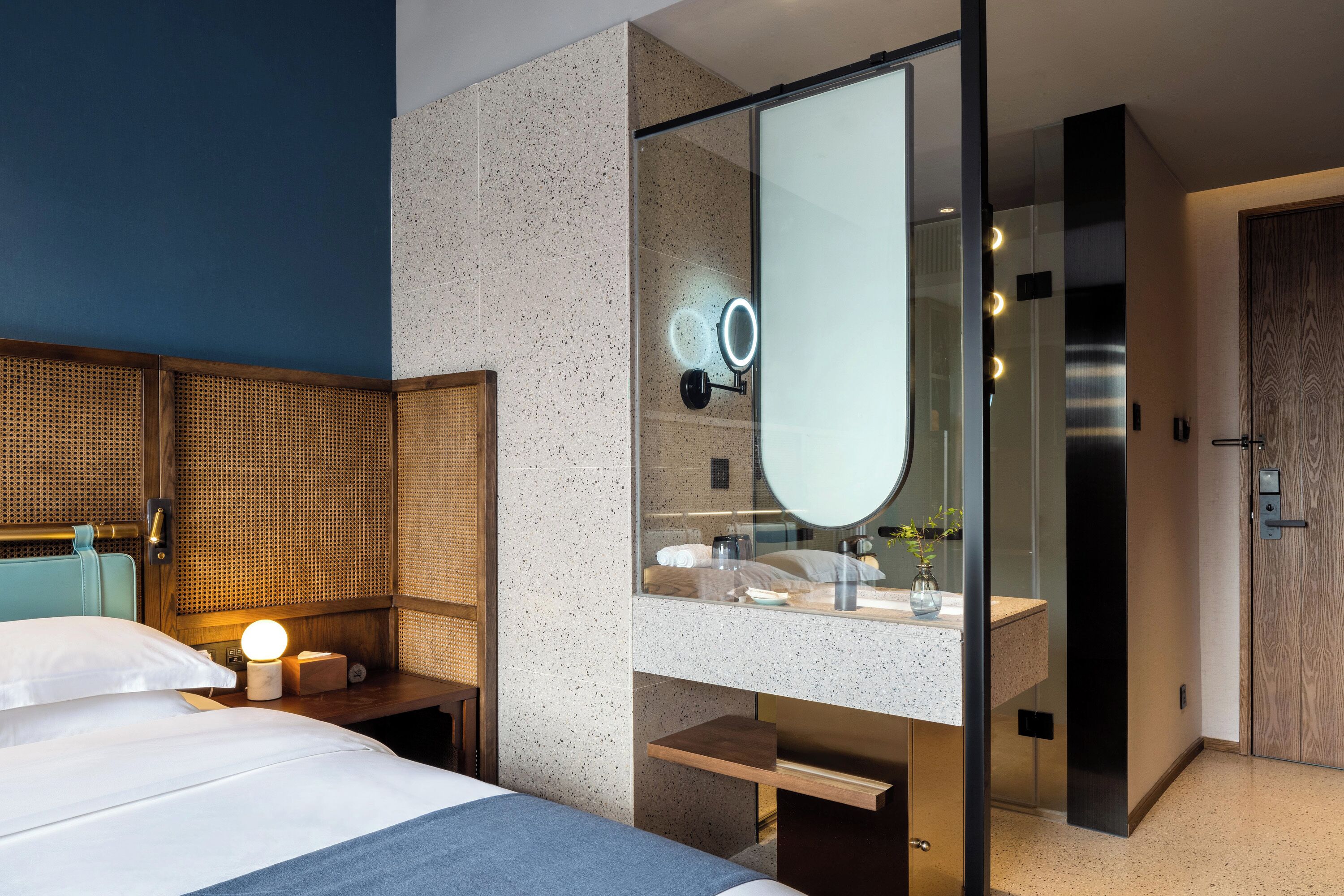 Quarto de hotel moderno com uma cama, mesa de cabeceira com abajur, espelho grande e banheiro com pia e chuveiro.