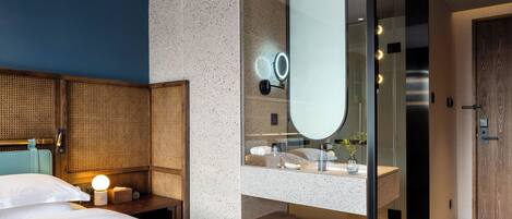 A modern hotel room with a bed, a bedside table with a lamp, a large mirror, and a bathroom with a sink and shower.