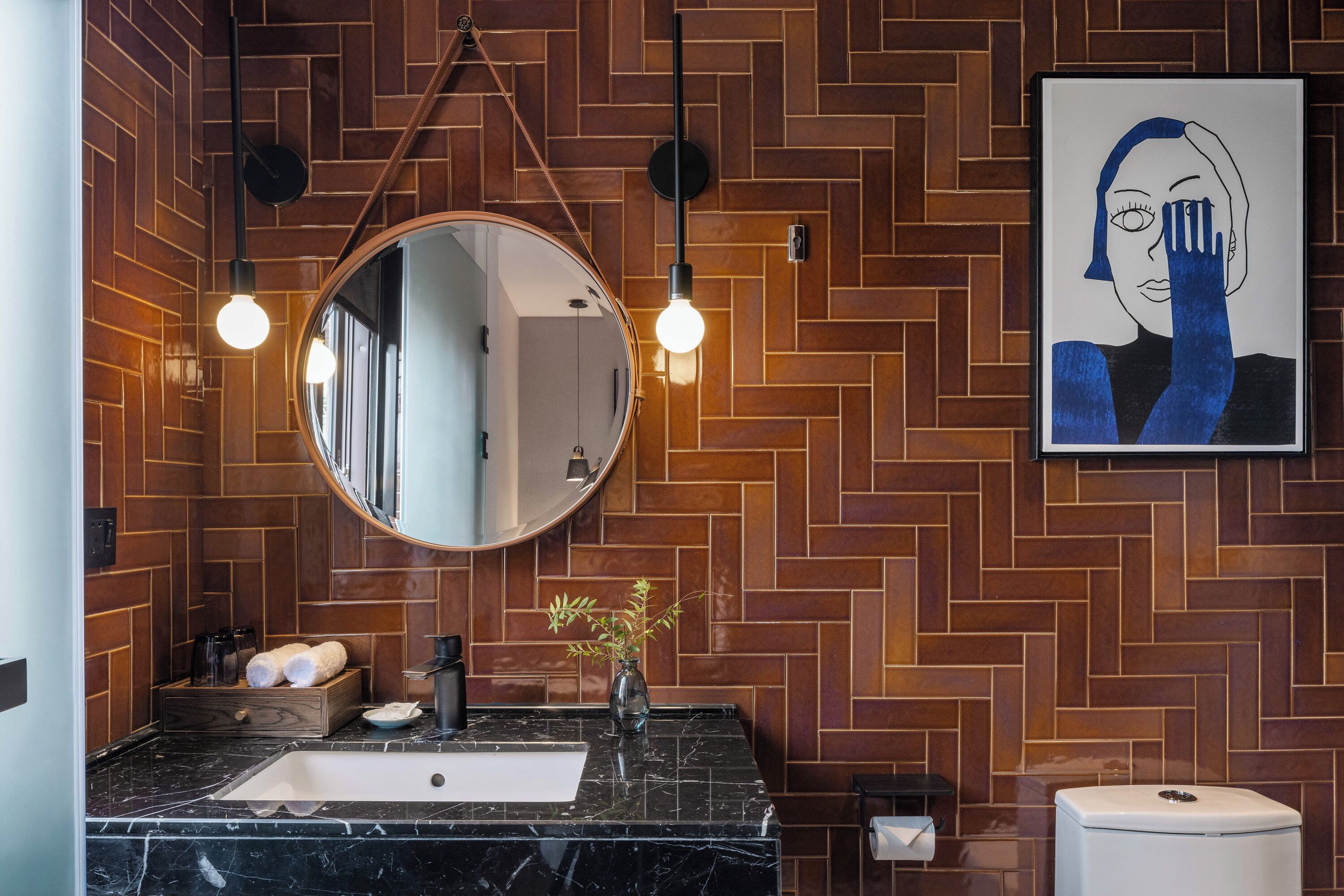 A bathroom with a herringbone-patterned wooden wall, a round mirror, a black marble sink, and a framed artwork.