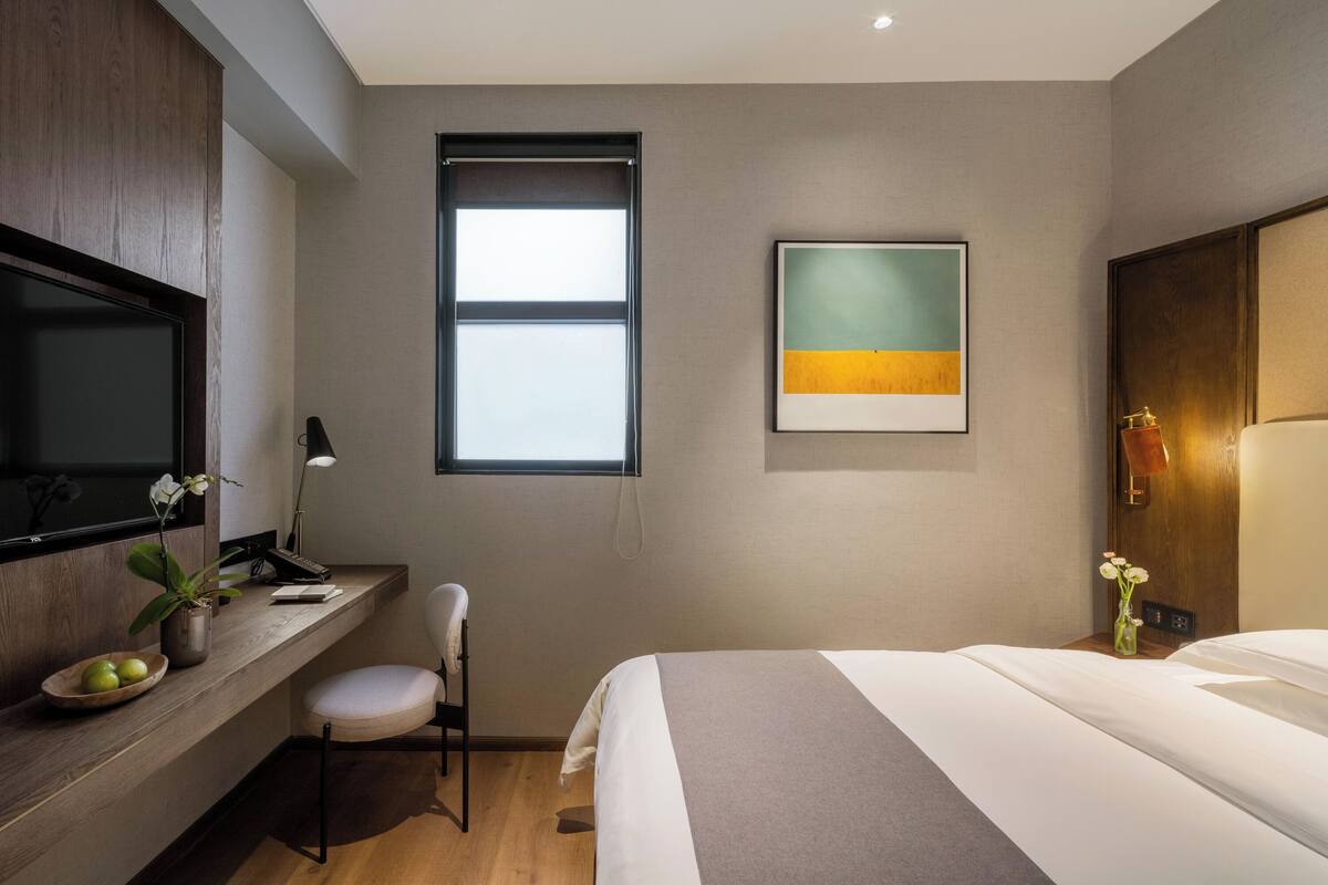 A modern hotel room with a bed, a desk with a chair, a television, a window, and a painting on the wall.