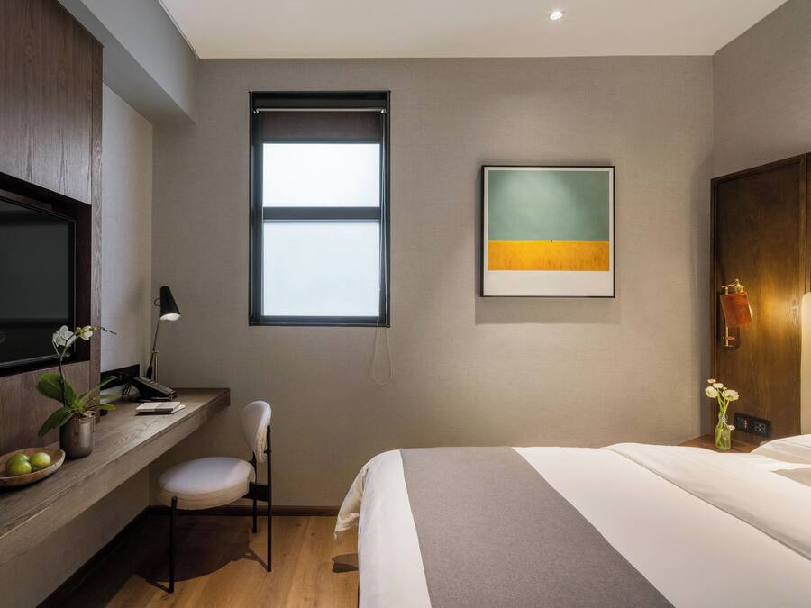 A modern hotel room with a bed, a desk with a chair, a television, a window, and a painting on the wall.