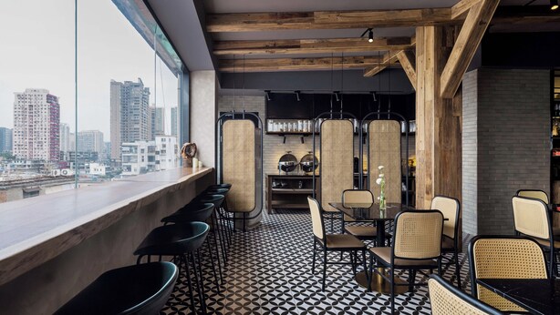 A modern interior with a city view, exposed wooden beams, black and white patterned flooring, and a mix of metal and wicker furniture.
