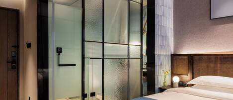 A modern hotel room with a large bed, a sliding glass door, a bedside table with a lamp, and a wall-mounted TV.