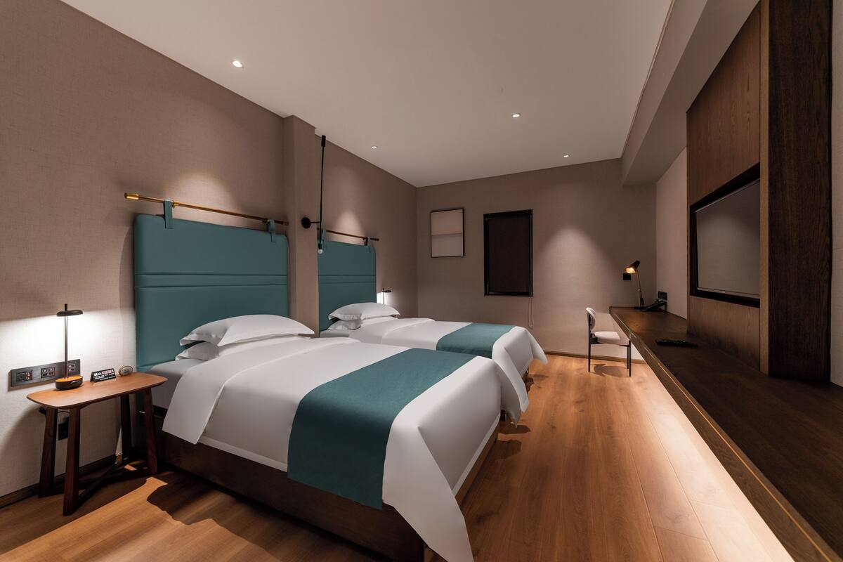 A modern hotel room with two beds, a wooden floor, a small desk, and a TV.