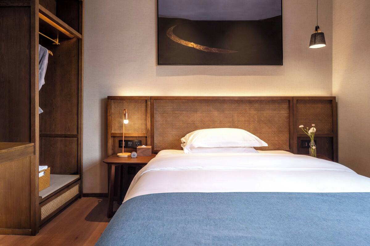 A hotel room with a large bed, a wooden wardrobe, a nightstand with a lamp, and a wall-mounted painting.