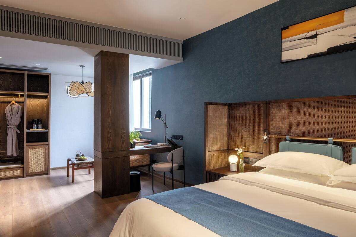 A modern hotel room with a large bed, a desk with a chair, a wardrobe, and a window providing natural light.