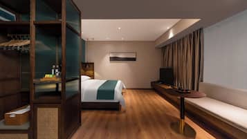 Una habitación de hotel moderna con suelo de madera, una cama con manta verde, un televisor en un mueble bajo, un sofá y un armario con espejo.