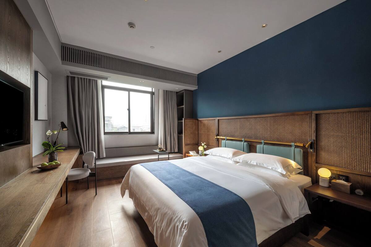 A modern hotel room with a large bed, a desk with a chair, a TV, and a window with curtains.