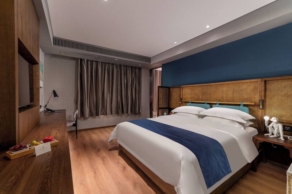 A modern hotel room with a large bed, wooden flooring, a desk with a lamp, and a small table with a decorative item.