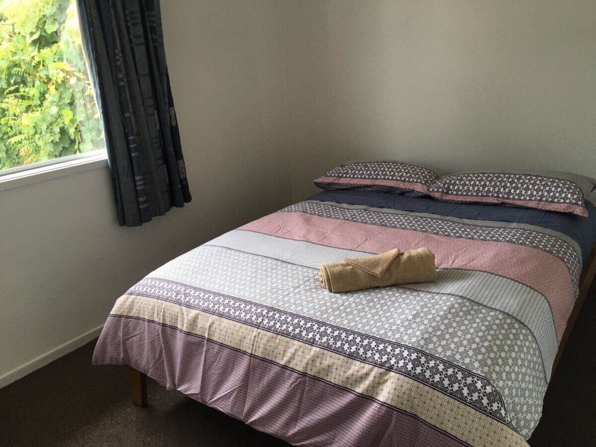 Cama bem arrumada com roupa de cama estampada e toalha enrolada, janela com cortinas e parede lisa.