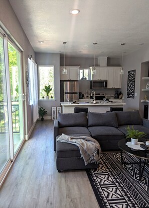 A modern living room with a sectional sofa, a dining area with a black table, and a kitchen with white cabinets and stainless steel appliances. - Midtown Midrise- Sapphire 200-The heart of Sacramento (Sacramento)