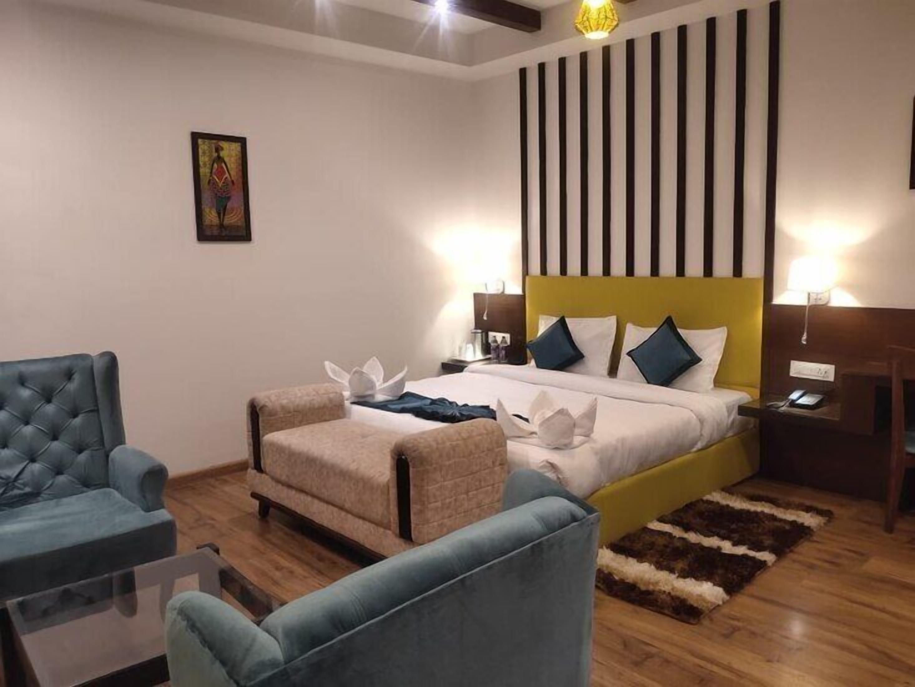 A hotel room with a large bed, two bedside tables, a blue armchair, a beige armchair, and a small table with a lamp.