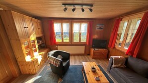 A cozy wooden cabin interior with a sofa, a black leather chair, a wooden table, and a television on a wooden cabinet.
