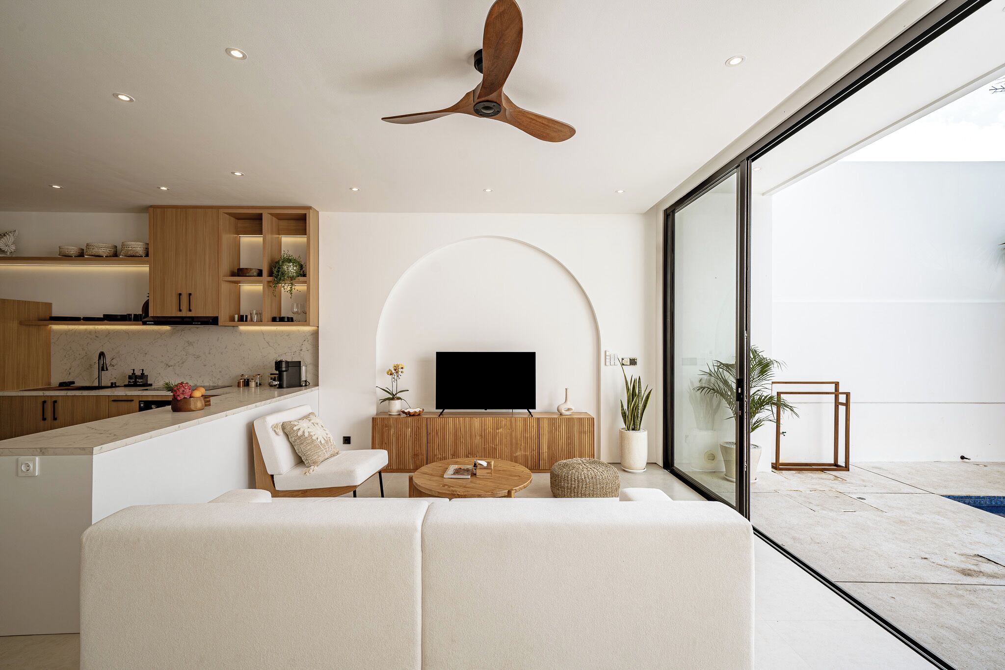 A modern living room with a sofa, coffee table, and TV, a kitchen with wooden cabinets, and a balcony with a pool.