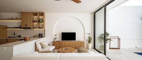 A modern living room with a sofa, coffee table, and TV, a kitchen with wooden cabinets, and a balcony with a pool.