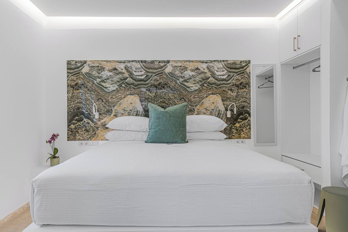 A modern bedroom with a large bed, a wall-mounted painting, a bedside table with a plant, and a wardrobe with a mirror.