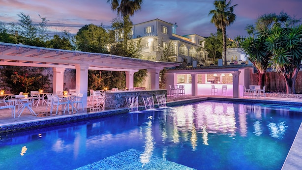 A pool area with a covered seating area, a bar, and a large building in the background. - Villa K Hotel (Corfu)